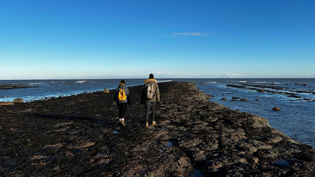 Two people walk along a narrow rocky spit of coast with sea either side