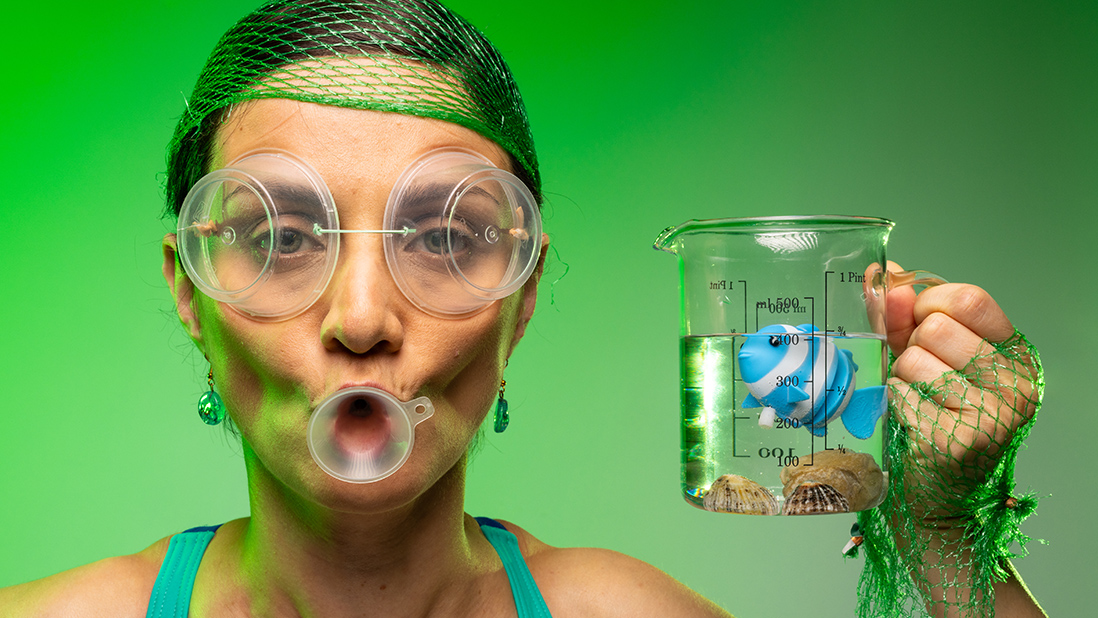 Artist Solmaz Farhang in goggles and a green net hat and green swimming costume holding a glass containing a blue fish